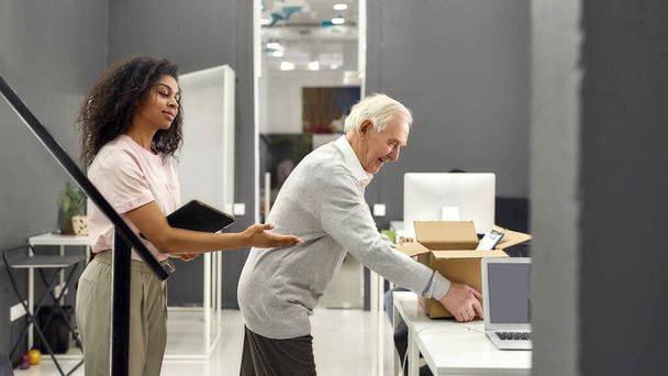 Friendly female company executive supporting, welcoming new employee aged man, senior intern putting cardboard box with belongings on the table - Photo, Image