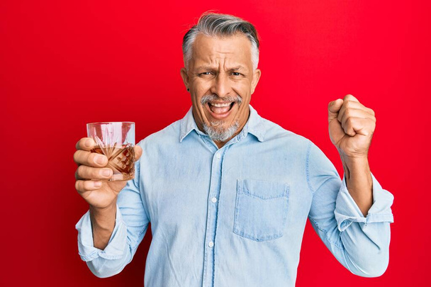Μεσαίωνας γκριζομάλλης άντρας πίνοντας ένα ποτήρι ουίσκι ουρλιάζοντας περήφανος, γιορτάζοντας τη νίκη και την επιτυχία πολύ ενθουσιασμένος με σηκωμένα χέρια  - Φωτογραφία, εικόνα