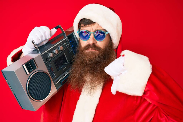 Handsome young red head man with long beard wearing santa claus costume and boombox with angry face, negative sign showing dislike with thumbs down, rejection concept  - Foto, Bild
