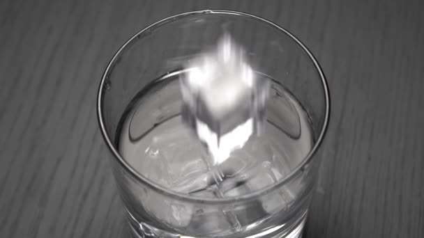 Langzame beweging van vallende ijsblokjes in water met spatten. Glas met transparante vloeistof op een zwarte grunge armoedige achtergrond. Close-up. - Video