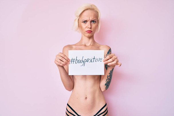 Молода блондинка з татуюванням, стоячи без сорочки, тримає банер з позитивним повідомленням тіла скептично і нервово, розчаровуючись через проблеми. негативна людина
.  - Фото, зображення