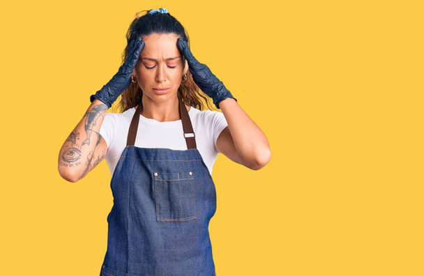 Jonge Spaanse vrouw met tatoeage met kappersschort en handschoenen die lijden aan hoofdpijn wanhopig en gestrest omdat pijn en migraine. handen op het hoofd.  - Foto, afbeelding