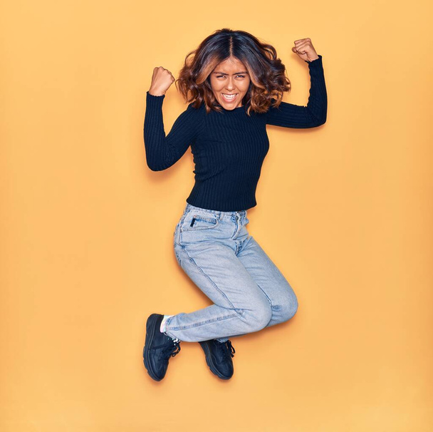 Jonge mooie latijnse vrouw die blij lacht. Springen met een glimlach op het gezicht vieren met vuisten omhoog over geïsoleerde gele achtergrond - Foto, afbeelding