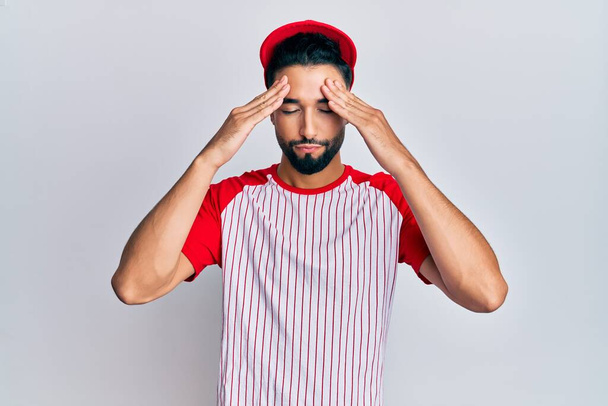 Молодий чоловік з бородою носить бейсбольну форму з рукою на голові, головний біль, тому що стрес. страждаюча мігрень
.  - Фото, зображення