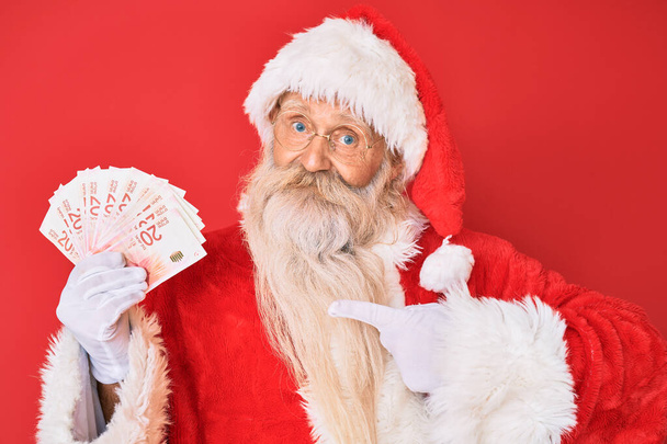 Staruszek z siwymi włosami i długą brodą w kostiumie Świętego Mikołaja trzymający izraelskie sykle uśmiechający się szczęśliwy wskazując ręką i palcem  - Zdjęcie, obraz