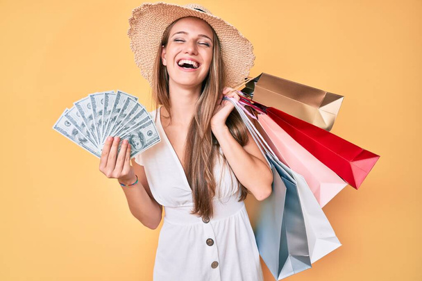 Young blonde girl holding shopping bags and dollars banknotes smiling and laughing hard out loud because funny crazy joke.  - 写真・画像