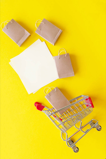 Mini carrello con piccole borse artigianali e carta bianca bianca bianca per lettering o shopping list su sfondo giallo. Il concetto di shopping nei negozi e su Internet. - Foto, immagini
