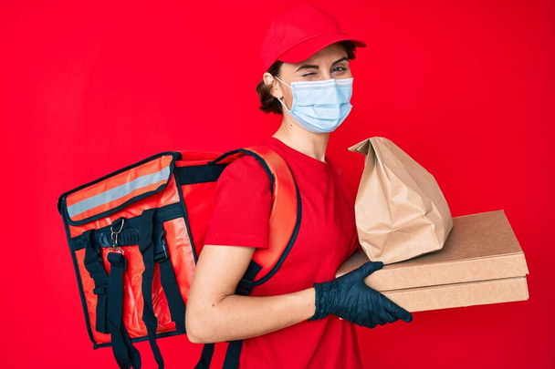 Young hispanic woman wearing medical mask holding take away food winking looking at the camera with sexy expression, cheerful and happy face.  - Photo, Image