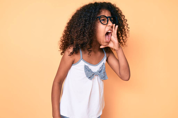カジュアルな服や眼鏡を身に着けている巻き毛を持つアフリカ系アメリカ人の子供は大声で叫び、口の上に手で横に大声で叫ぶ。コミュニケーションの概念.  - 写真・画像