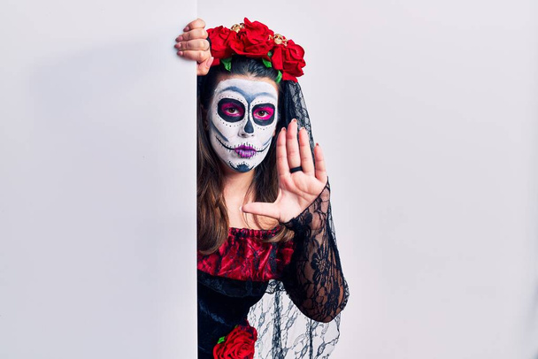 Jonge vrouw draagt dag van de dode custome houden lege banner doen stoppen met zingen met palm van de hand. waarschuwingsuitdrukking met negatief en ernstig gebaar op het gezicht.  - Foto, afbeelding
