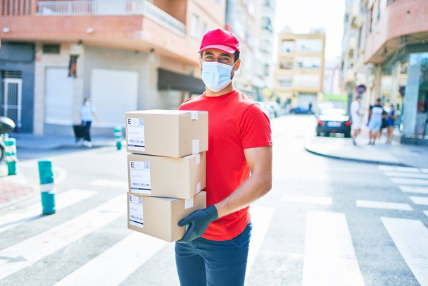 Junger Zusteller in Uniform und Coronavirus-Schutzmaske hält Pappschachteln an der Stadtstraße. - Foto, Bild