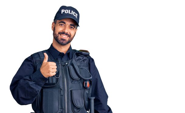 Junger hispanischer Mann in Polizeiuniform macht glückliche Daumen-hoch-Gesten mit der Hand. Zustimmender Blick in die Kamera zeigt Erfolg.  - Foto, Bild