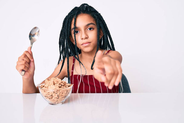 Niña afroamericana joven con trenzas sentada en la mesa comiendo cereales señalando con el dedo a la cámara y a ti, gesto confiado que se ve serio  - Foto, imagen