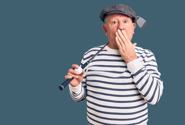 Starszy przystojny siwowłosy mężczyzna trzymający kij golfowy i piłkę zakrywającą usta ręką, zszokowany i bojący się pomyłki. zaskoczona ekspresja  - Zdjęcie, obraz