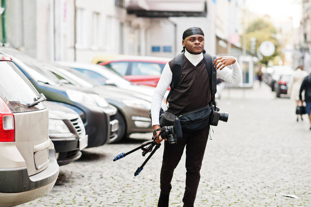 Young professional african american videographer holding professional camera with tripod pro equipment. Afro cameraman wearing black duraq making a videos. - Foto, Imagem
