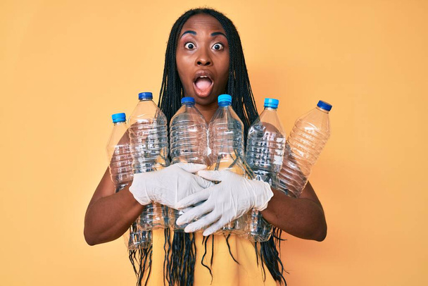 Afro-Amerikaanse vrouw met vlechten houden recycling plastic flessen bang en geschokt met verrassing en verbaasd uitdrukking, angst en opgewonden gezicht.  - Foto, afbeelding