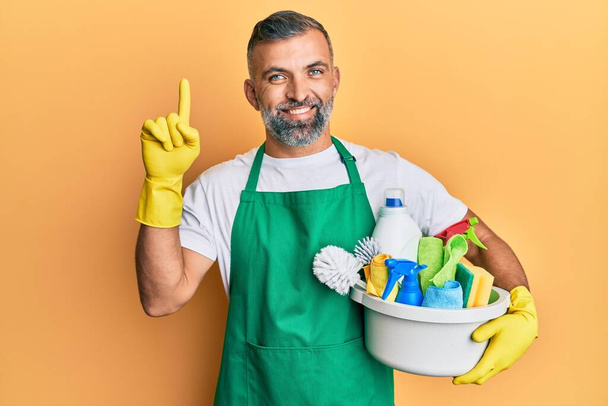 Homem bonito de meia idade segurando produtos de limpeza surpreso com uma ideia ou pergunta apontando o dedo com o rosto feliz, número um  - Foto, Imagem