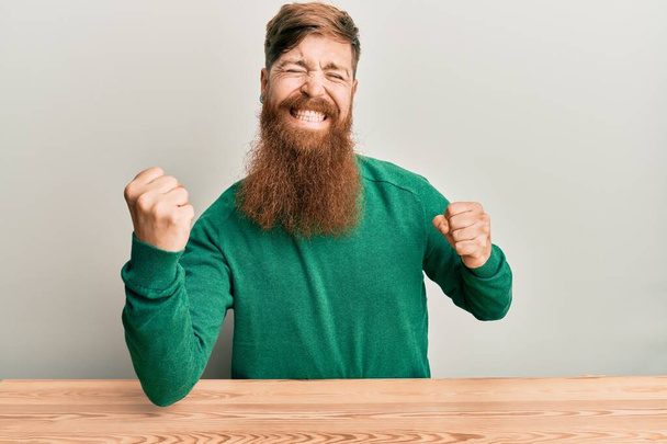 Jonge Ierse roodharige man in casual kleding op de tafel zit erg blij en enthousiast doen winnaar gebaar met opgeheven armen, glimlachen en schreeuwen om succes. viering concept.  - Foto, afbeelding