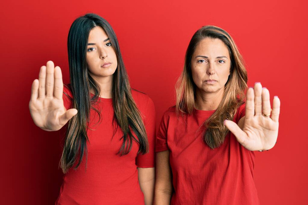 赤い背景の上にカジュアルな服を着ている母親と娘のヒスパニック系の家族は、手の手のひらで歌うのを停止します。顔に負の重大なジェスチャーで.  - 写真・画像
