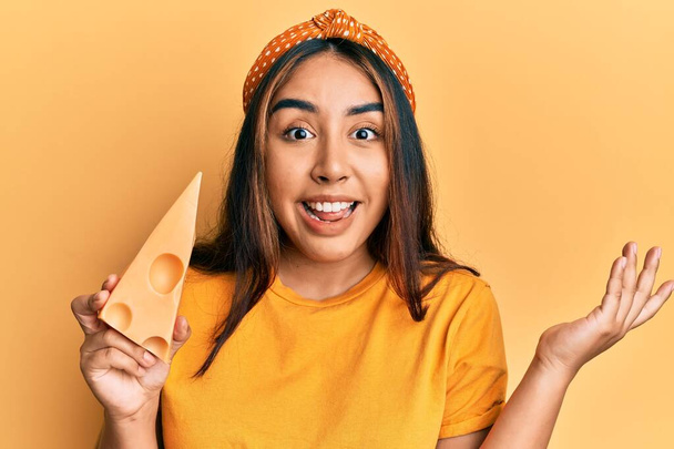 Молодая латинская женщина ест сыр Эмменталь празднуя достижение с счастливой улыбкой и выражение лица победителя с поднятой рукой  - Фото, изображение