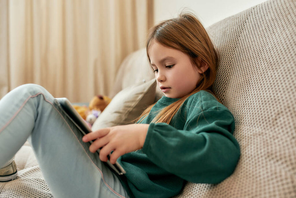 Egy aranyos kislány, aki a táblagépével van elfoglalva, miközben a kanapén ül. - Fotó, kép