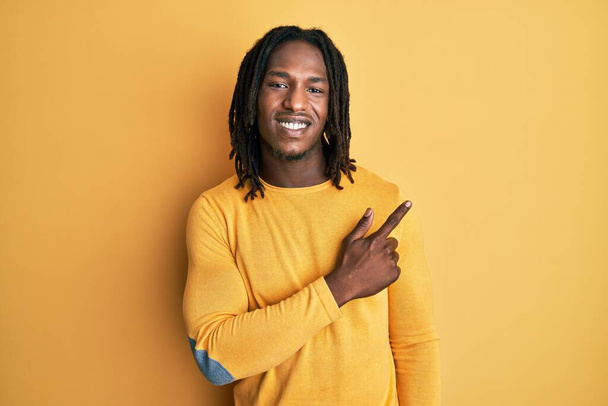 Afrykański Amerykanin z warkoczami w żółtym swetrze, uśmiechnięty, pogodny, wskazujący ręką i palcem z boku.  - Zdjęcie, obraz