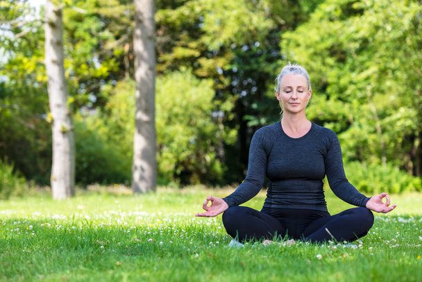 Mature middle aged fit healthy woman practicing yoga outsidein a natural tranquil green environment - Photo, image