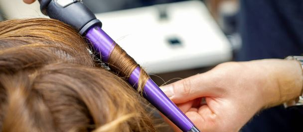 Professzionális fodrász teszi göndör frizura curling vas hosszú vörös haj a fiatal nő a fodrászatban - Fotó, kép