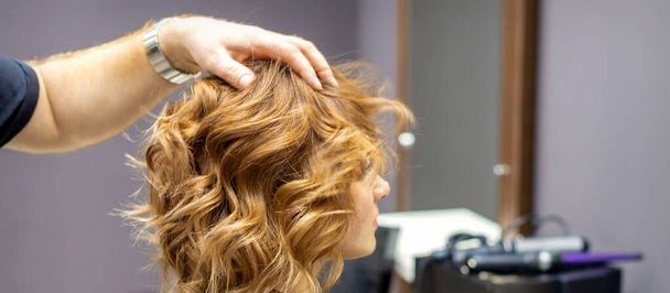 Seitenansicht des männlichen Friseurs überprüft lockige Frisur der jungen kaukasischen Frau im Friseursalon - Foto, Bild
