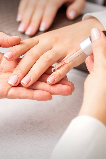 Hand van manicure giet olie door pipet aan nagelriemen van nagels van jonge vrouw in schoonheidssalon. Franse manicure - Foto, afbeelding