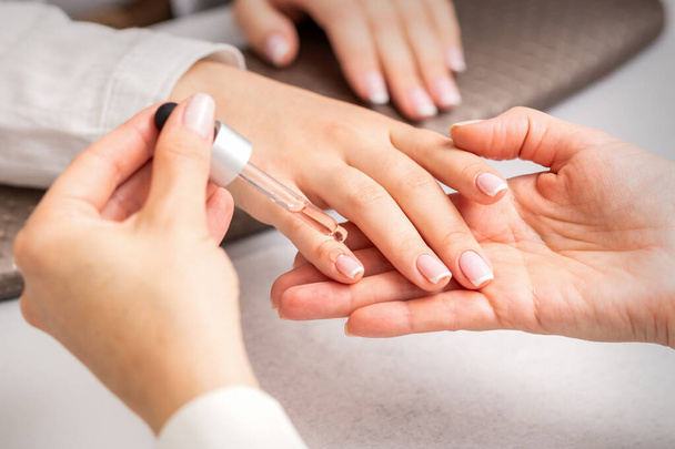Hand of manicurist pours oil by pipette to cuticle of nails of young woman in beauty salon. French manicure - 写真・画像