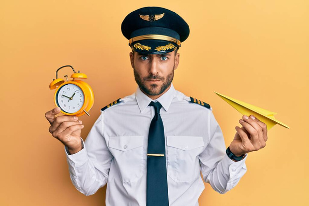 Handsome hispanic pilot man holding paper plane and alarm clock skeptic and nervous, frowning upset because of problem. negative person.  - Φωτογραφία, εικόνα