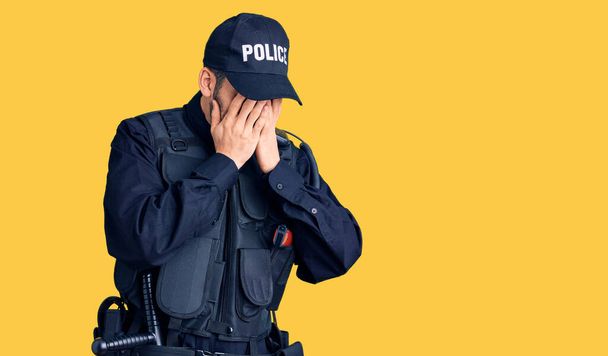 Giovane bell'uomo con la barba che indossa l'uniforme della polizia con espressione triste che copre il viso con le mani mentre piange. concetto di depressione.  - Foto, immagini