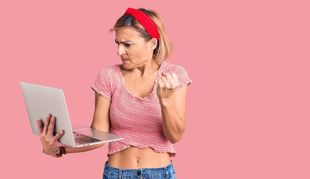 Jeune femme blonde tenant un ordinateur portable ennuyé et frustré criant de colère, criant fou de colère et la main levée  - Photo, image