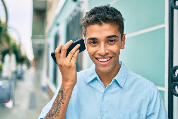 Genç Latin adam gülümsüyor. Şehirde akıllı telefon kullanarak mutlu bir sesli mesaj dinliyor.. - Fotoğraf, Görsel