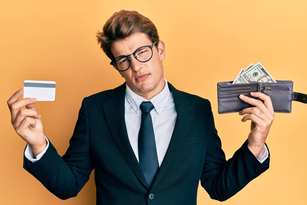 Handsome caucasian business man holding credit card and wallet with dollars clueless and confused expression. doubt concept.  - Fotoğraf, Görsel