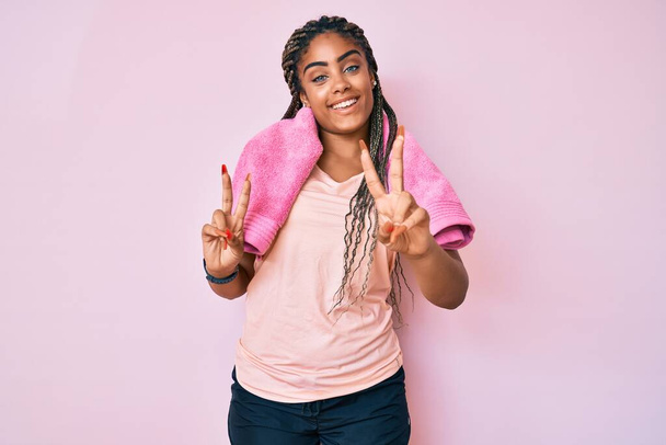 Fiatal afro-amerikai nő copfban, sportruhában és törülközőben, mosolyogva nézi a kamerát, amint ujjakat mutat győzelmi jelekkel. Második..  - Fotó, kép