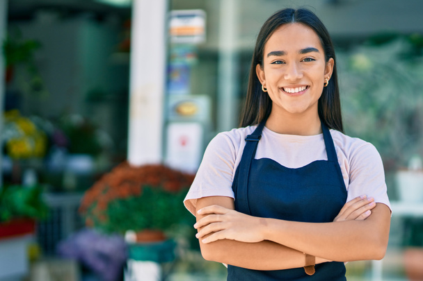 Young latin shopkeeper girl with arms crossed smiling happy standing at the florist - Photo, Image