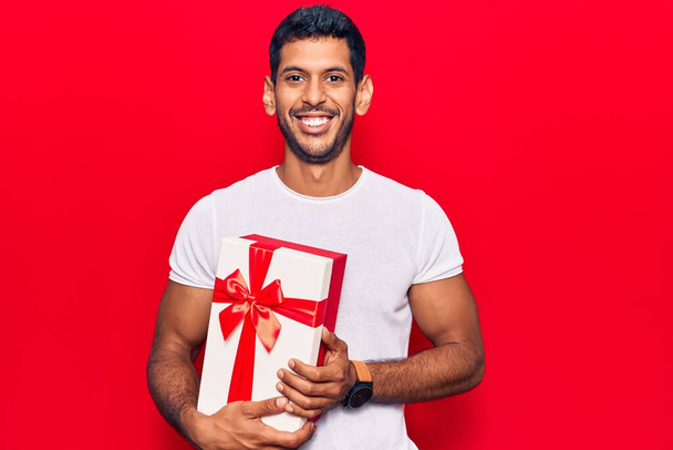 Young latin man holding gift looking positive and happy standing and smiling with a confident smile showing teeth  - Foto, imagen