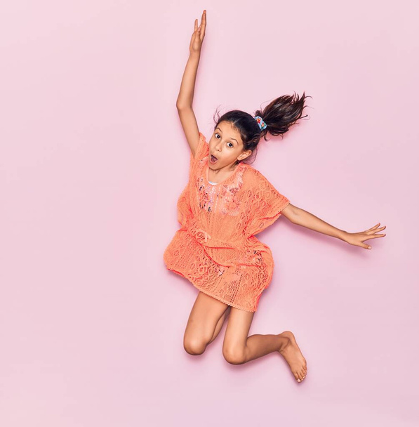 Αξιολάτρευτο ισπανόφωνο κορίτσι σε διακοπές φορώντας καλοκαιρινό φόρεμα χαμογελώντας ευτυχισμένη. Άλμα με χαμόγελο στο πρόσωπο πάνω από απομονωμένο ροζ φόντο - Φωτογραφία, εικόνα