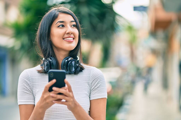 Joven mujer hispana sonriendo feliz usando teléfonos inteligentes y auriculares en la ciudad. - Foto, imagen