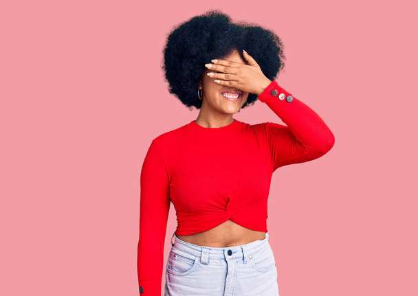 Молодая африканская американская девушка в повседневной одежде и очках улыбается и смеется, закрывая глаза рукой на лице для сюрприза. слепая концепция.  - Фото, изображение