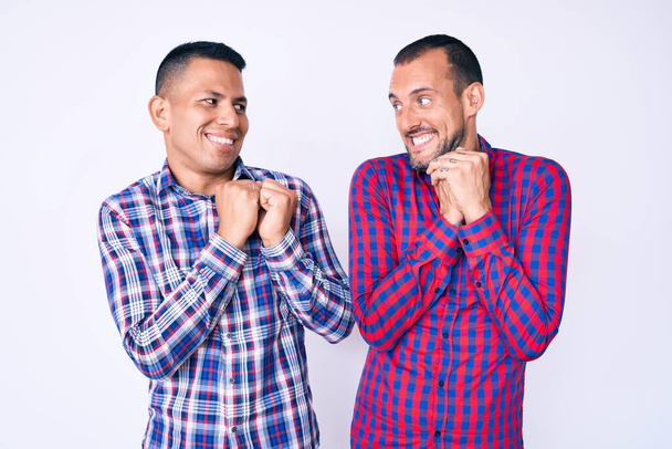 jong gay paar van twee mannen dragen casual kleding lachen nerveus en enthousiast met handen op kin kijken naar de zijkant  - Foto, afbeelding