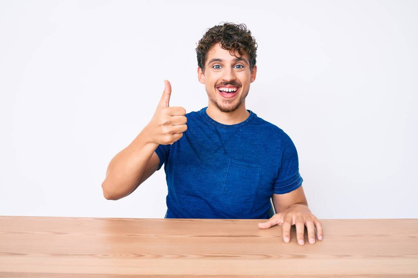 Junger kaukasischer Mann mit lockigem Haar, lässig gekleidet auf dem Tisch sitzend, glücklich und positiv lächelnd, Daumen hoch und Zustimmungszeichen  - Foto, Bild