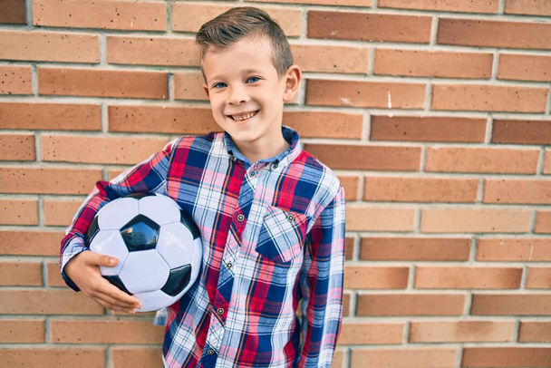Adorable chico caucásico sonriendo feliz sosteniendo pelota de fútbol de pie en la ciudad - Foto, Imagen
