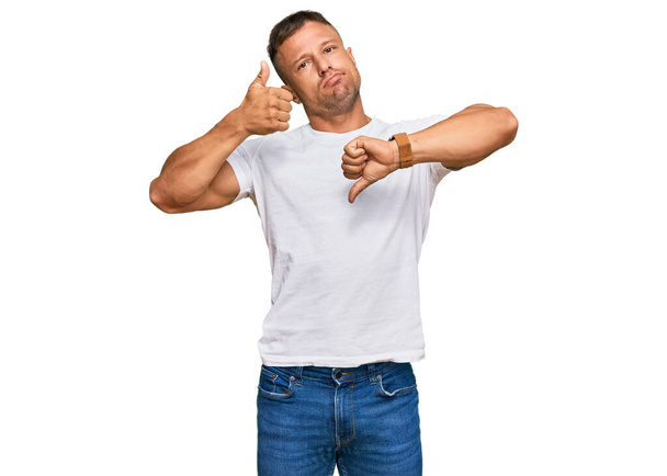 Hombre musculoso guapo usando camiseta blanca casual haciendo pulgares arriba y abajo, desacuerdo y expresión de acuerdo. conflicto loco  - Foto, Imagen