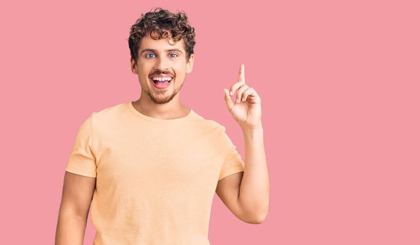 Young handsome man with curly hair wearing casual clothes pointing finger up with successful idea. exited and happy. number one.  - Photo, Image
