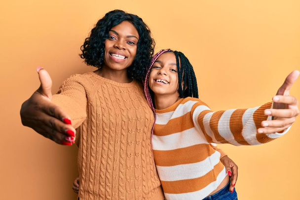 Schöne afrikanisch-amerikanische Mutter und Tochter tragen wollenen Winterpullover und blicken lächelnd mit offenen Armen in die Kamera, um sich zu umarmen. fröhlicher Ausdruck, der das Glück umarmt.  - Foto, Bild