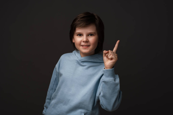Ciddi bir çocuk portresi. 10-12 yaşlarında, günlük giysiler içinde. İşaret parmağı gösteriyor, azarlıyor ya da azarlıyor. Küçük çocuk parmağını kaldırdı ve "Dikkat lütfen" dedi. Stüdyo çekimi, gri arkaplan - Fotoğraf, Görsel