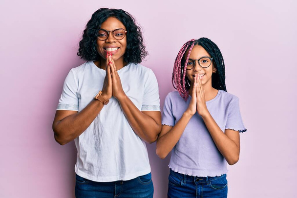Όμορφη αφροαμερικανή μητέρα και κόρη φορώντας καθημερινά ρούχα και γυαλιά προσεύχονται με τα χέρια μαζί ζητώντας συγχώρεση χαμογελώντας σίγουροι.  - Φωτογραφία, εικόνα